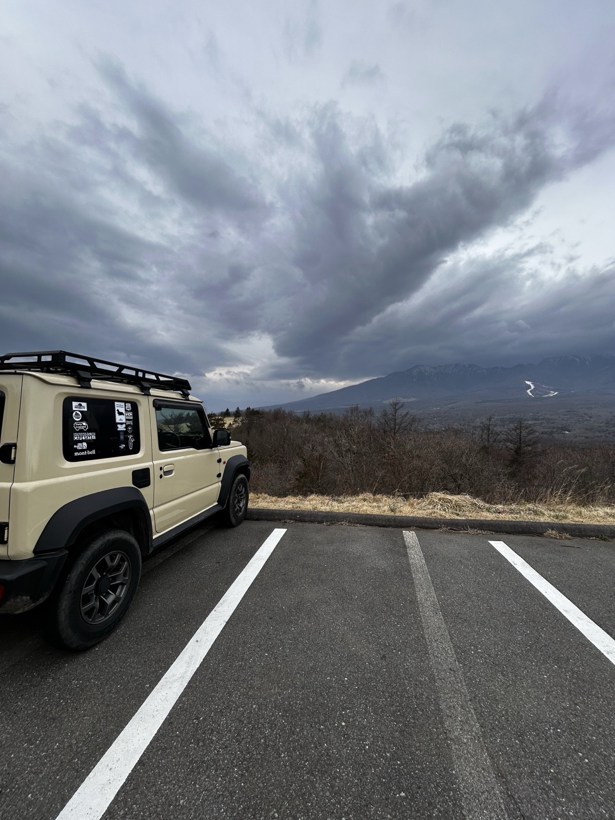 山をバックに駐車場に停まるジムニーシエラ(JB74)。背面タイヤとステッカーが見える